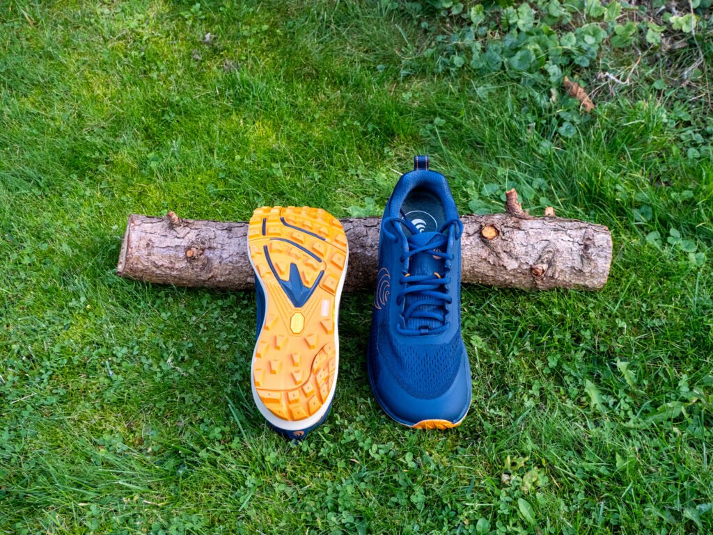 Topo Athletic Terraventure 5 mit oranger Vibram-Megagrip-Sohle neben dem Schuh von vorn